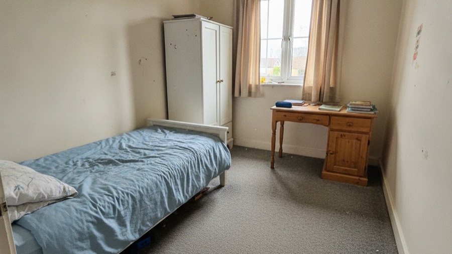 A kids room before redesign, with a plain single bed, scuffed pine desk and worn grey carpet